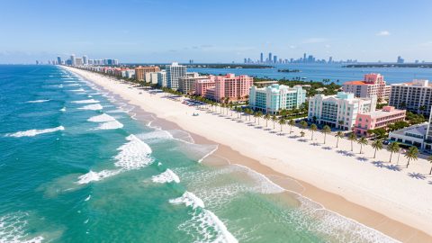 Vue aérienne lumineuse de Miami Beach avec mer turquoise, palmiers, hôtels Art déco pastel et skyline moderne au loin.