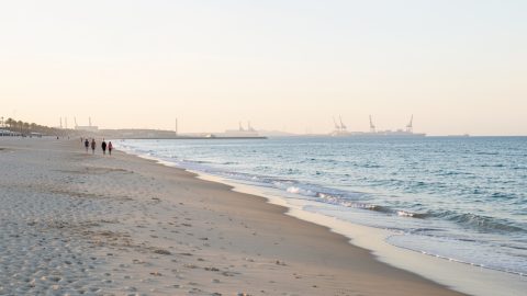 Vue panoramique réaliste de la Grande Plage de Fos-sur-Mer au matin, avec du sable clair, la Méditerranée bleue, quelques promeneurs éloignés et la silhouette du port à l’horizon.