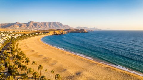 Vue aérienne lumineuse de la baie d’Agadir en octobre, avec une grande plage de sable doré, l’océan Atlantique, des palmiers et les montagnes de l’Anti-Atlas à l’horizon.