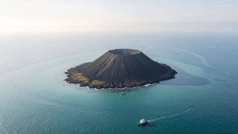 Vue aérienne réaliste du Krakatoa, île volcanique sombre entourée d’eau turquoise dans le détroit de la Sonde, avec un petit bateau d’excursion au loin sous la lumière matinale.
