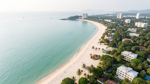 Vue aérienne réaliste de la plage de Wong Amat à Pattaya, avec son sable clair, ses hôtels discrets, sa végétation tropicale et la mer calme du golfe de Thaïlande baignée par la lumière matinale.