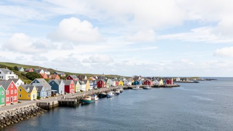 Vue panoramique du port de Saint-Pierre-et-Miquelon avec des maisons colorées au bord de l’eau, des bateaux et un ciel lumineux.